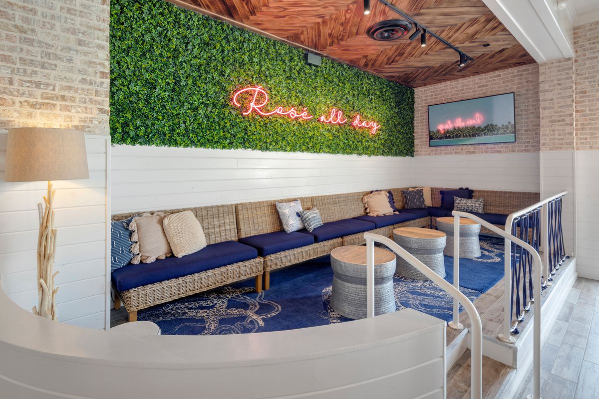A blue and white beach-themed lounge area with a faux ivy wall with a neon pink sign above the wicker couch reading, “rose all day” at the Hampton Social. 