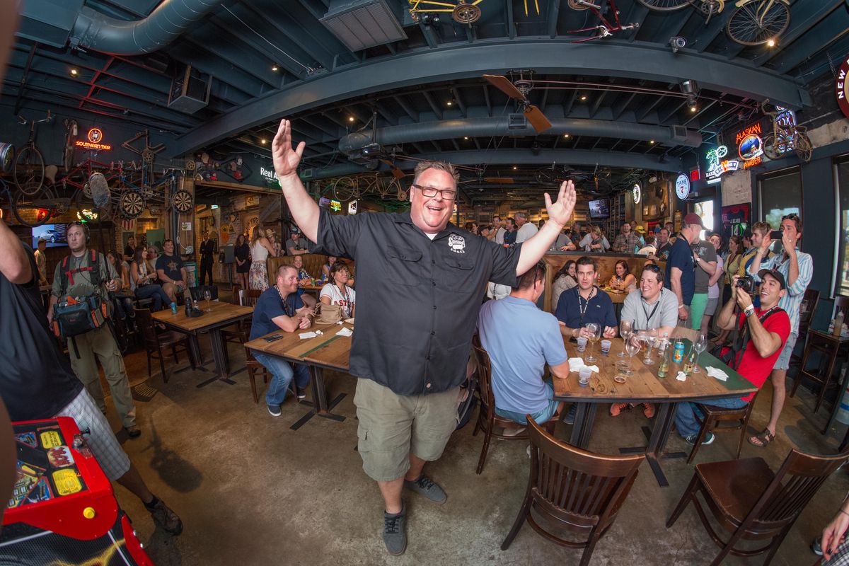 Chef Chris Shepherd cheerfully raising his hands in the air in front of a crowded bar.
