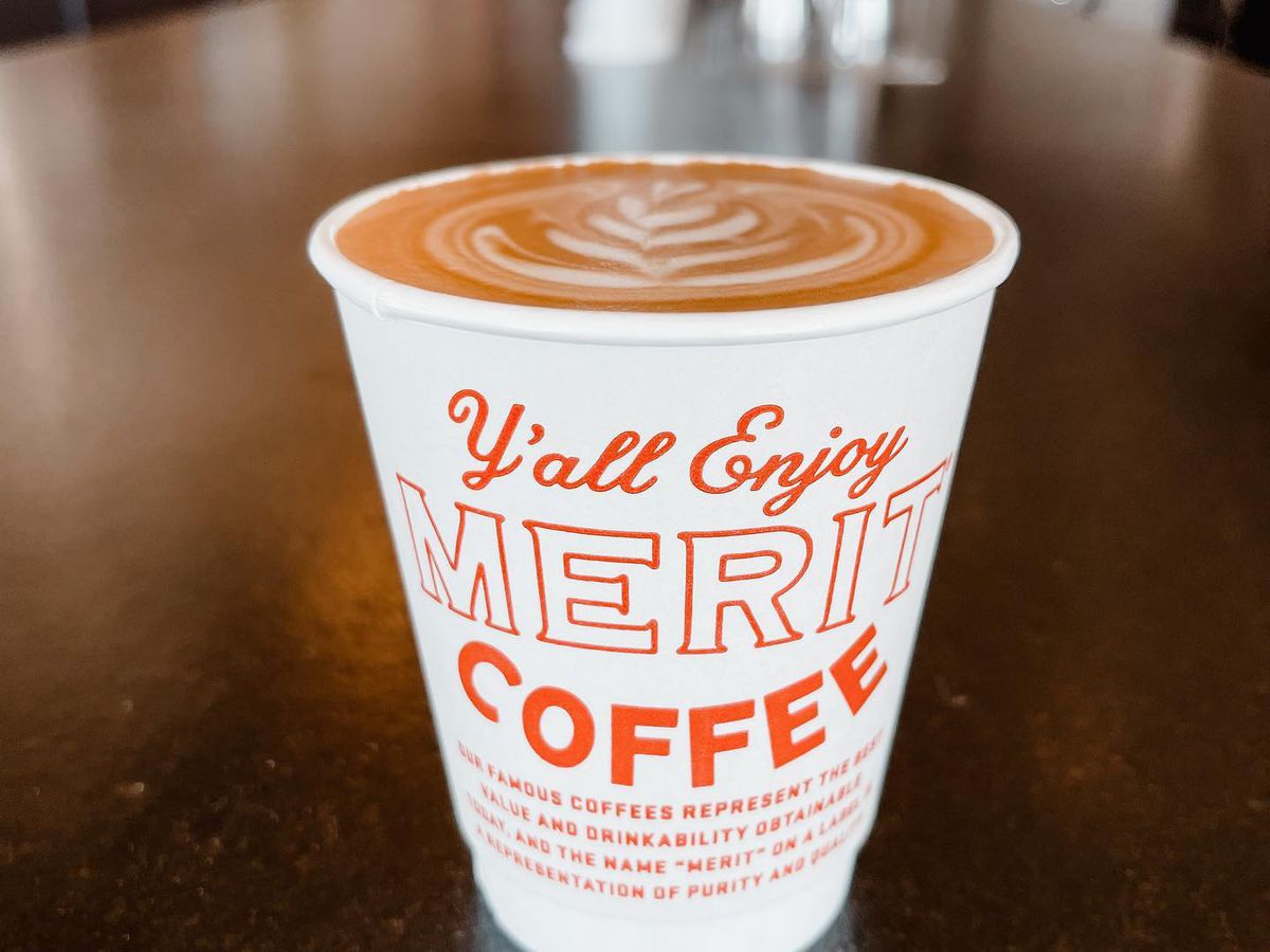 Closeup of a coffee in a white coffee cup with “Y’all enjoy Merit Coffee” printed on it in red letters. 