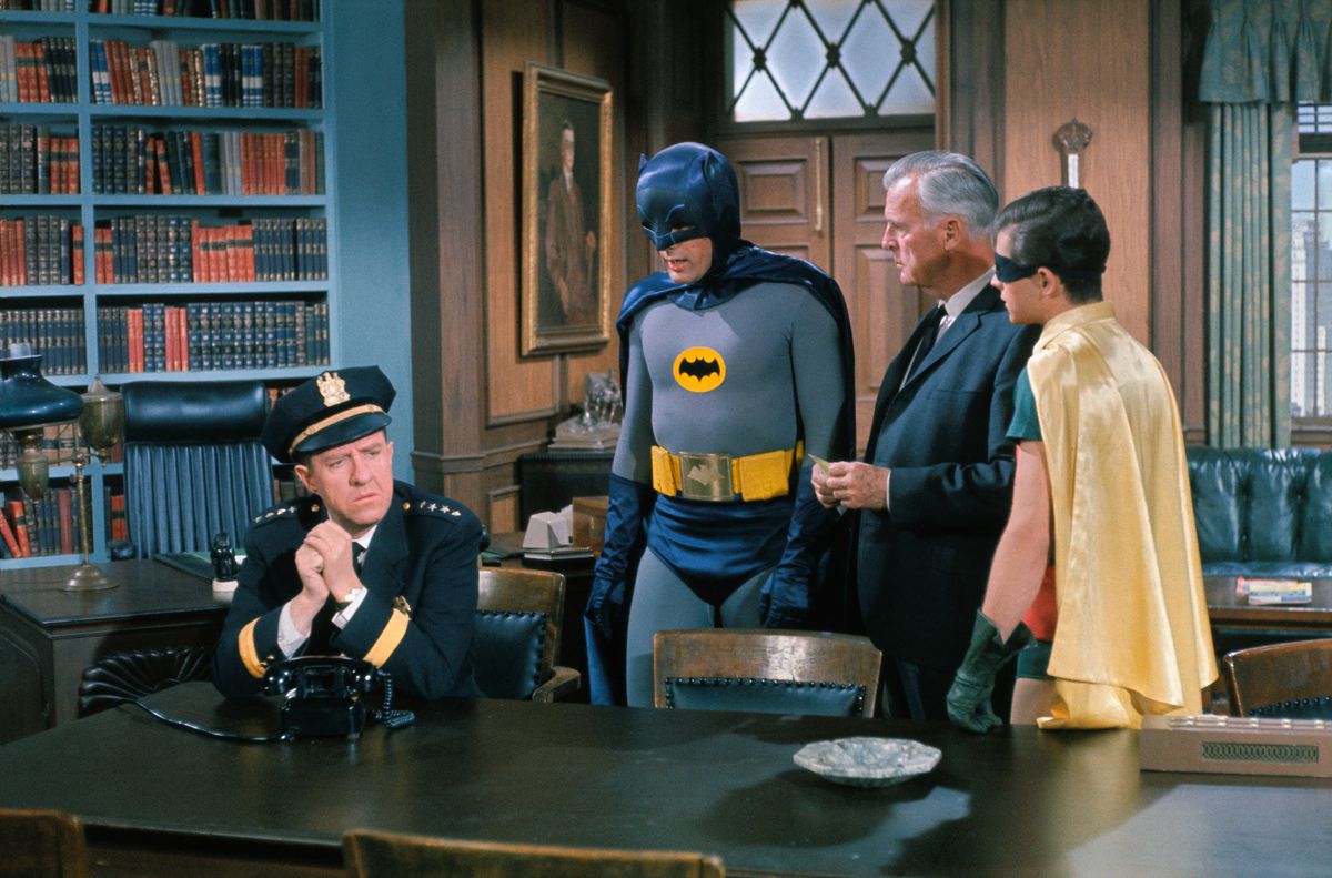Adam West and Burt Ward as Batman and Robin, stand in a library talking with Commissioner Gordon and Chief O’Hara in Batman (1966).