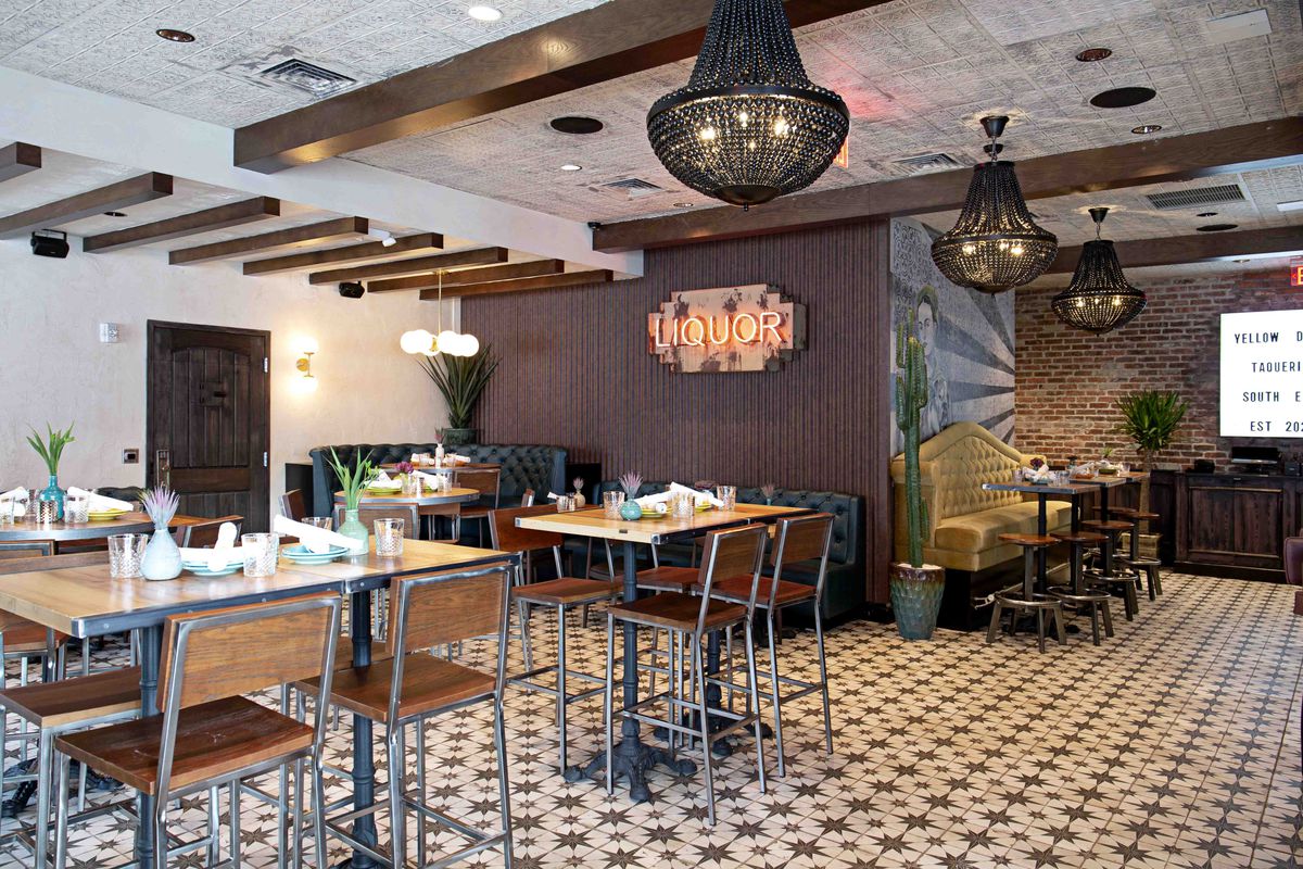 Wide-angle view of a restaurant shows patterned tile flooring, wooden tables and chairs with metal supports, dark beams in the ceiling, exposed brick walls, and neon accent signs