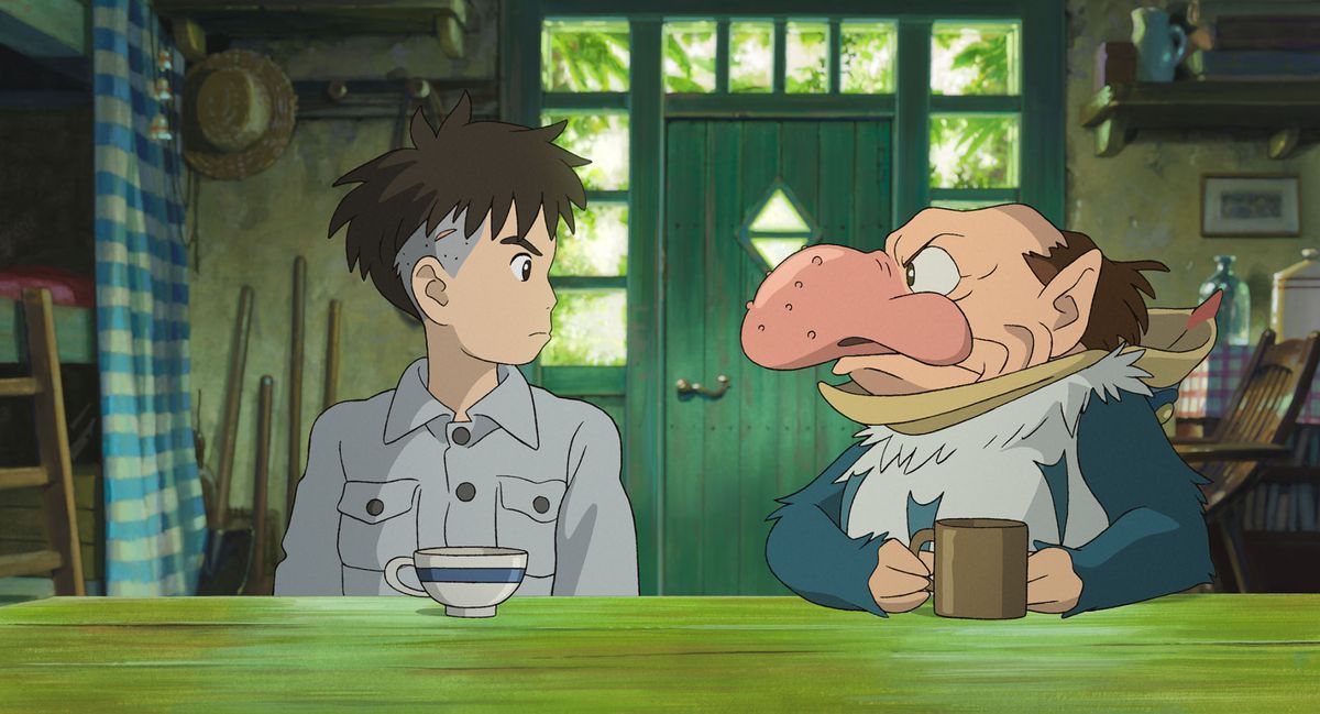 A stubborn-looking boy with shaggy hair and an undercut sits across the table from a small man with a huge, pimple-covered nose and a bald head with a brown fringe around his pointed ears, wearing a blue-and-white feathered suit and clutching a brown mug, in Hayao Miyazaki’s The Boy and the Heron
