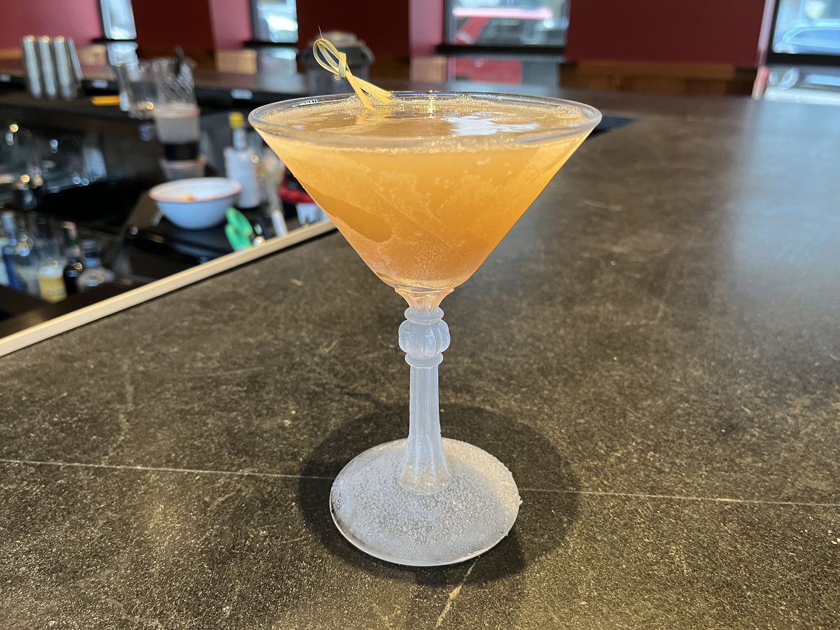 A long stemmed martini glass with orange liquid inside, garnished with a fruit peel.