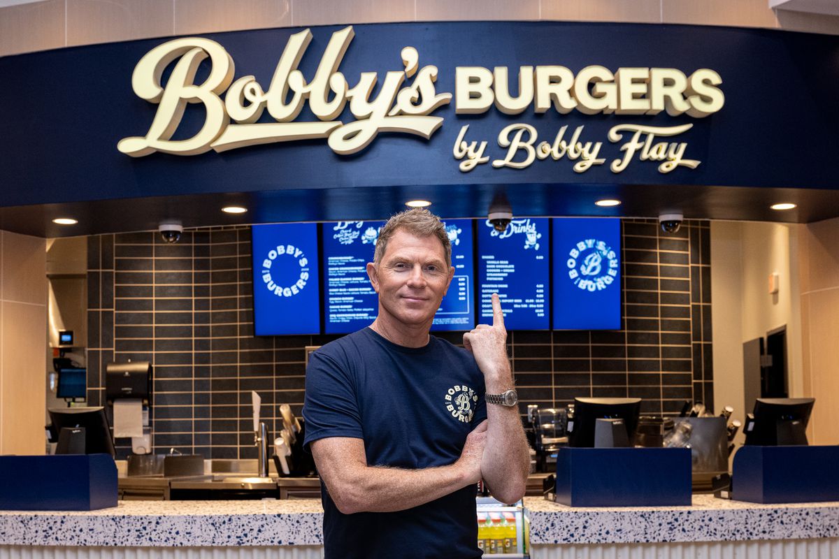Celebrity Bobbly Flay points to a sign that reads, “Bobby’s Burgers.” 