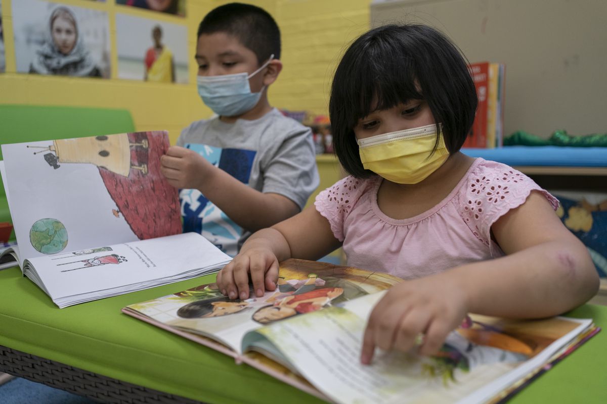 A young boy wearing a blue face mask and a gray shirt looks at a book. Next to him a young girl with a yellow face mask and a pink shirt looks at another book. Both are at a lime green desk or table.