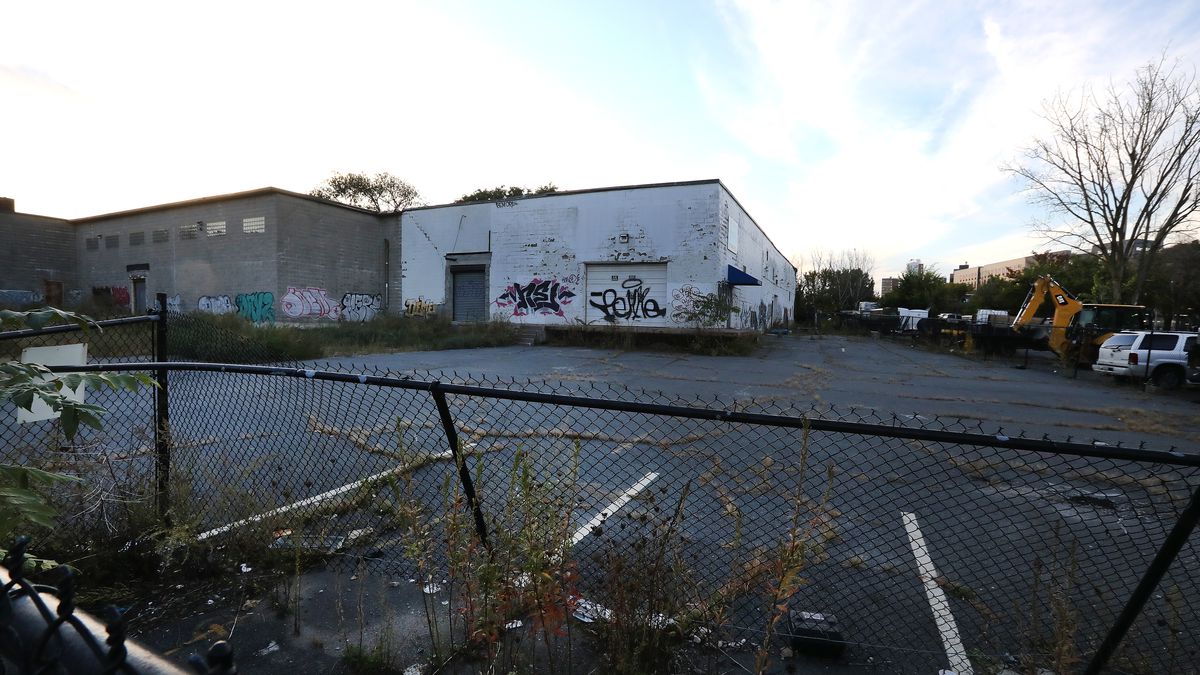 A vacant lot with a fence in front of it and a rundown single-story building at the end of it. 