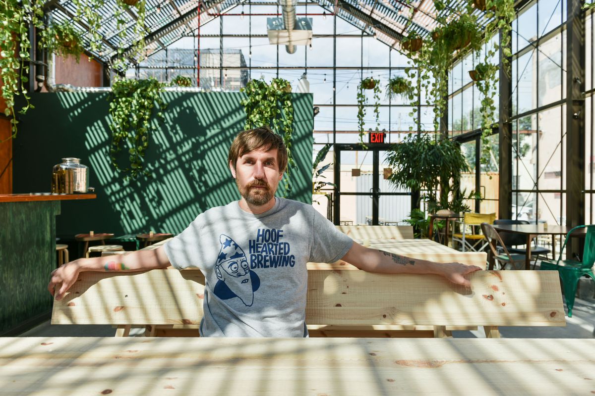 Jeppe Jarnit-Bjergsø sitting at a wooden table inside a greenhouse with plants behind him