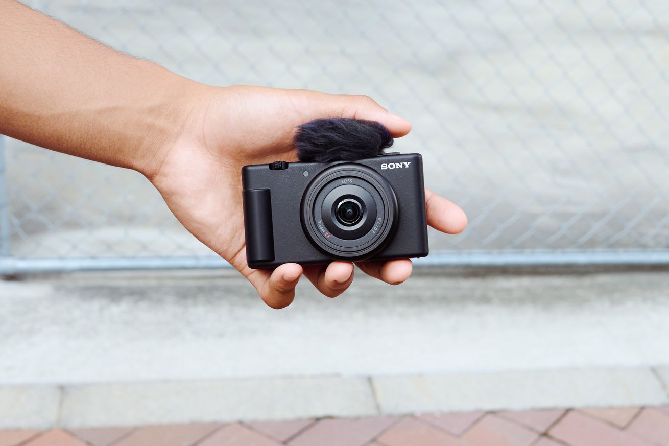 Front view of Sony’s black ZV-1F compact camera with lens cap removed and wind screen attached, held in a person’s hand in front of a wall.