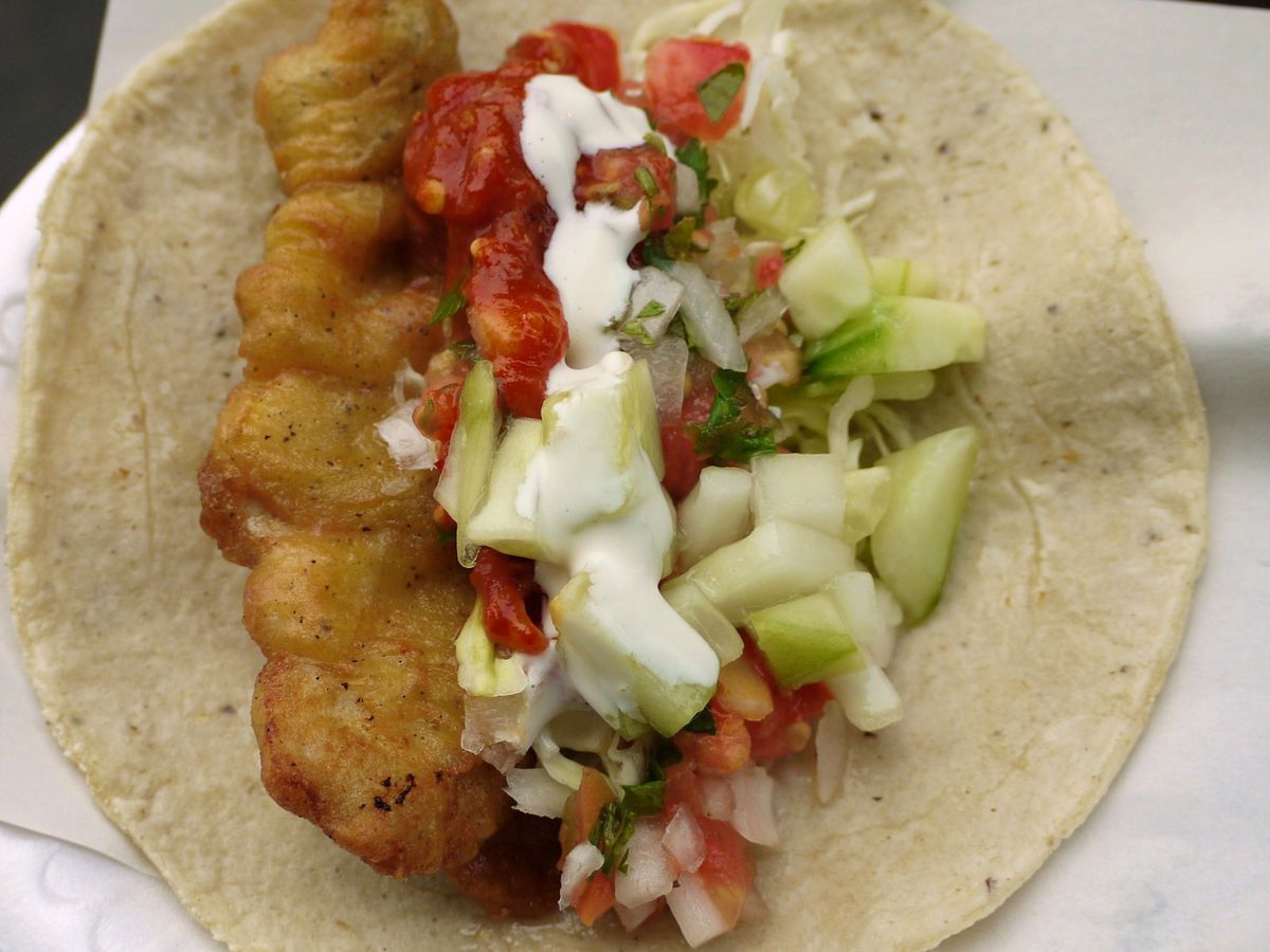 From above, A taco with a long strip of fried fish on one side, beside a long mound of chopped vegetables and multiple sauces