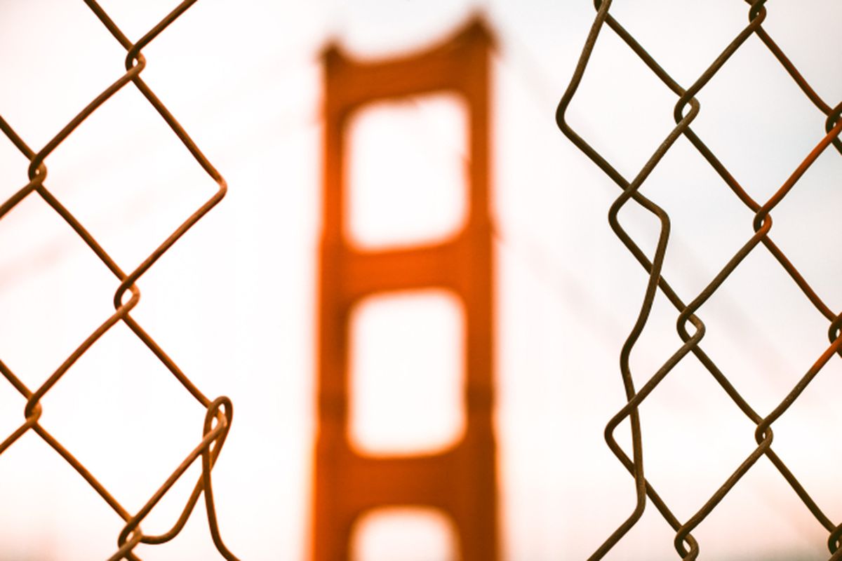 An orange-painted tower of a suspension bridge, seen through a whole in a chainlink fence.
