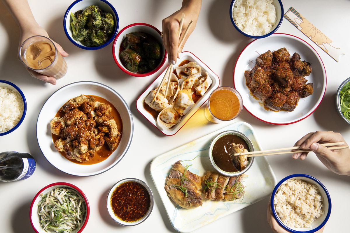 A series of bowls with Chinese food like dumplings, roasted duck, and spicy cauliflower on a white table with some hands holding chopsticks.
