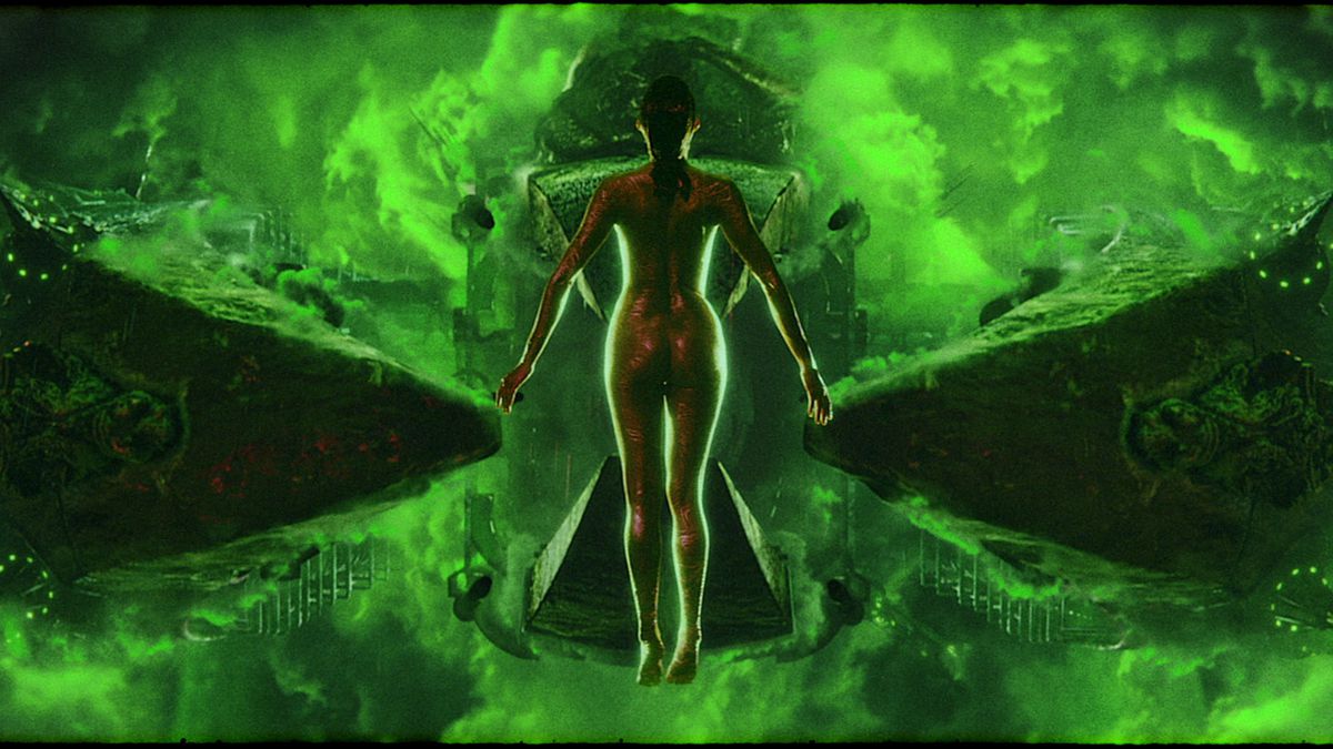 A naked female form, back to the camera, hangs in space in front of a glowing green hourglass shape made out of light