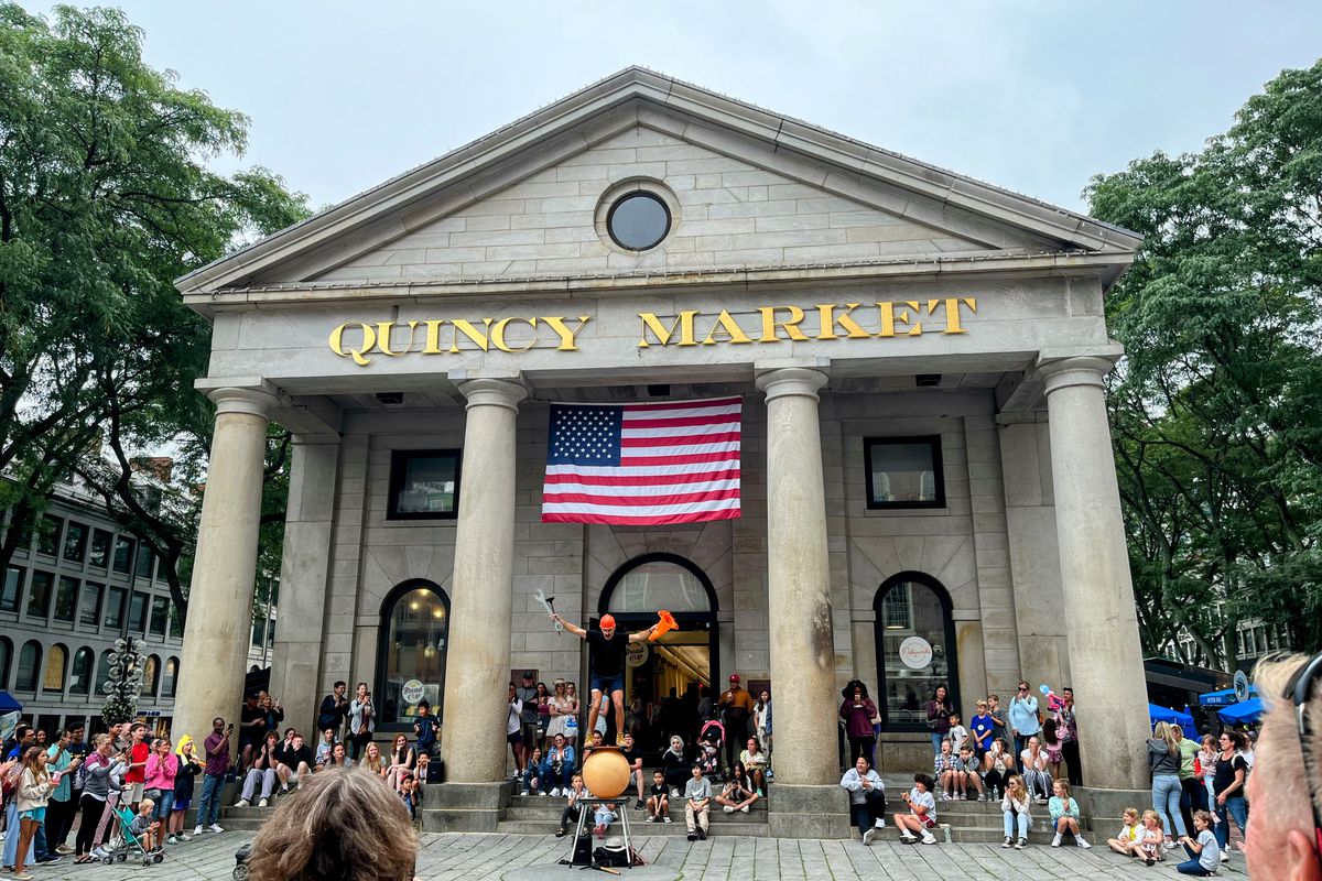 A juggler balances on a large orange ball in front of a crowd outside of Quincy Market in downtown Boston, performing tricks.
