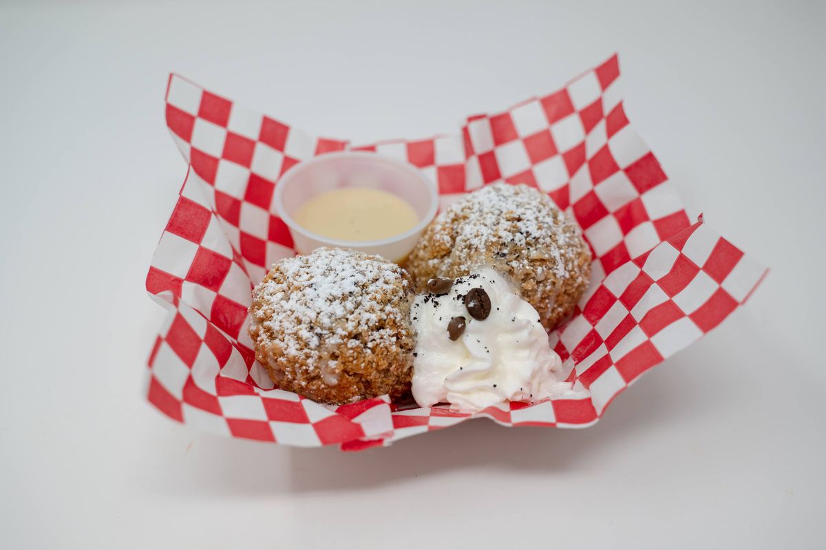 Two fried balls sit in a boat on a red-checked paper. In front of them is a dollop of whipped cream with coffee beans. Behind is a plastic container of dolce de leche.