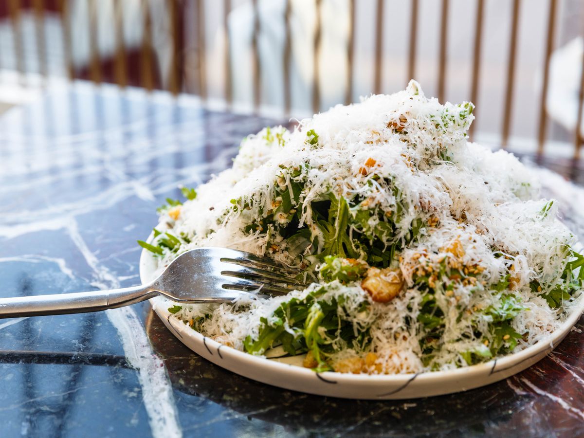 A green salad showered in cheese shavings with a fork placed to the side of the dish sits on a table.