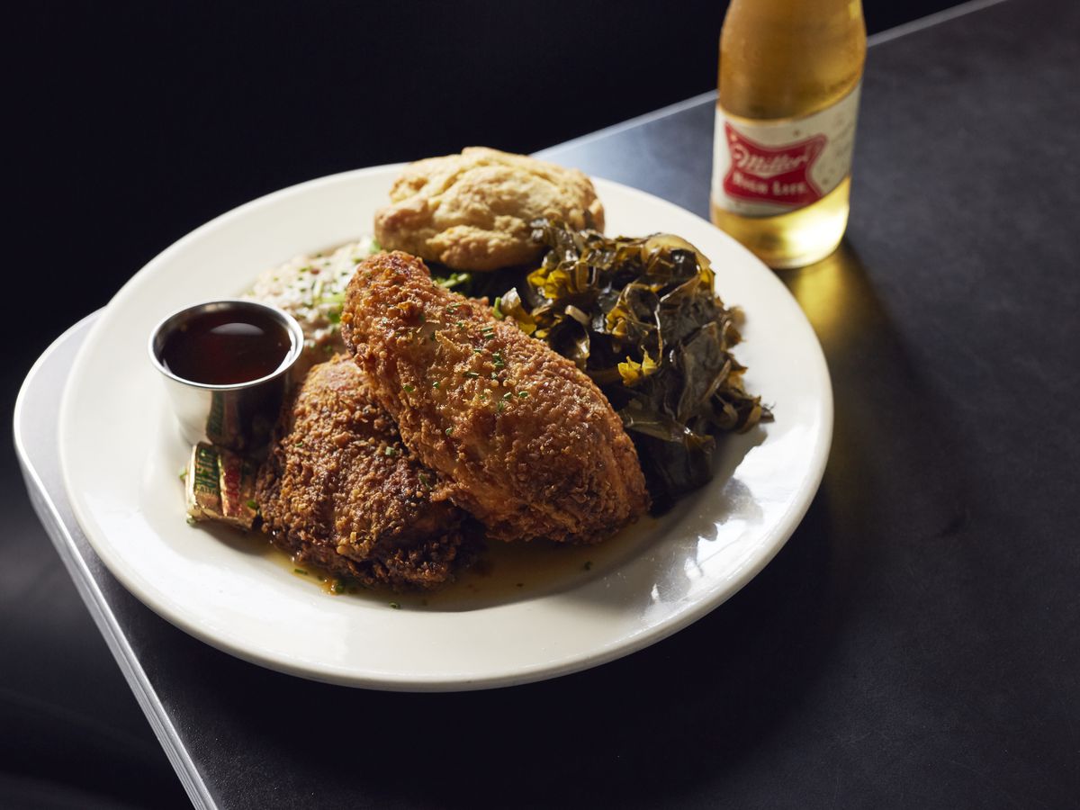 A plate of fried chicken with collard greens and a biscuit sits on a black tabletop on a black background. There’s a bottle of Miller High Life to the side of the plate.