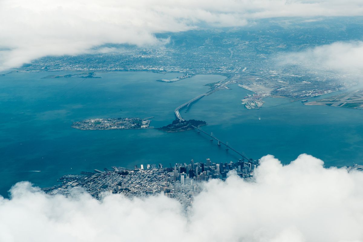 An aerial photo of an island surrounded by blue bay waters on all sides, with bridges connecting to dense cities on the north and south sides and clouds in the foreground.