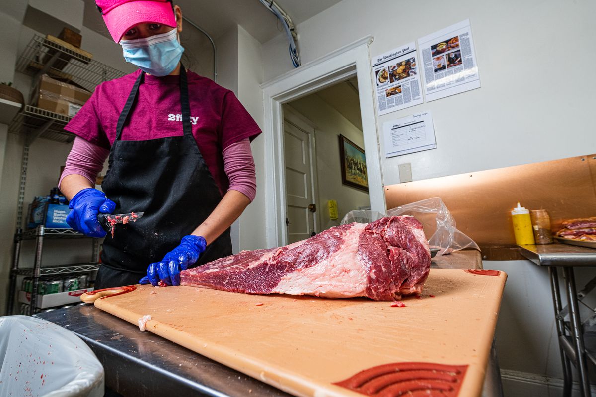 A 2Fifty employee wearing a branded magenta shirt and holding a knife to trim a slab of brisket on a cutting board.