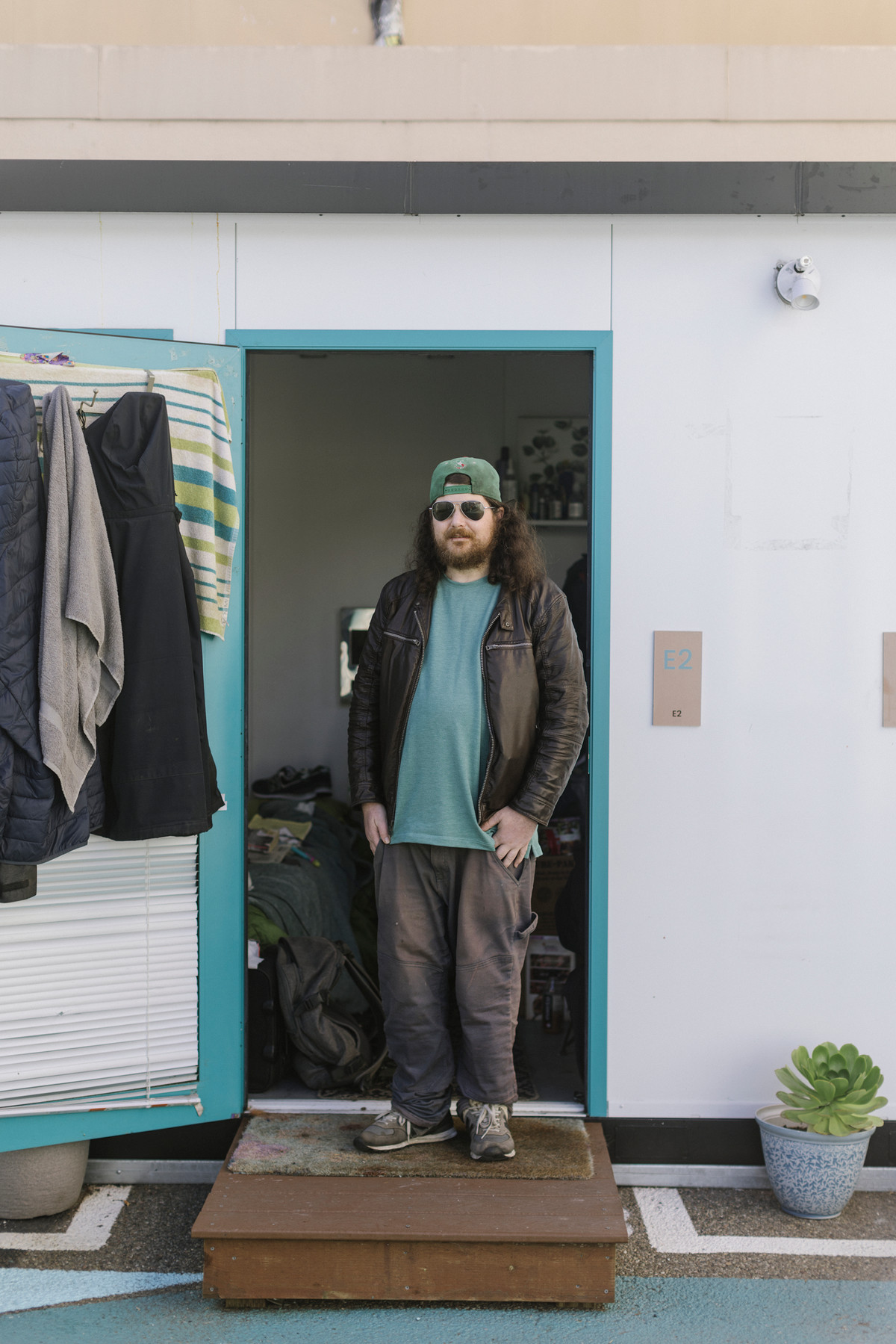 A man with long dark hair, a cap, and glasses, stands in the doorway of a tiny house.