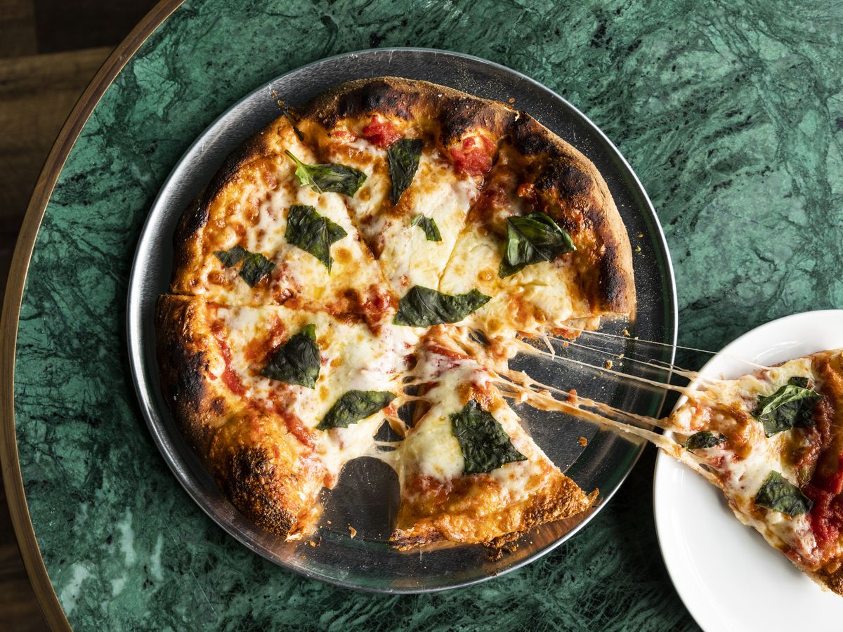 A piece of pizza lies on a plate with cheese strings stretching from the center of the pie. The plate sits on a green marble table.
