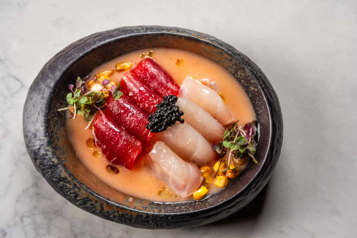 Rolls of raw fish sit in a tan broth inside of a stone bowl on a marble table.