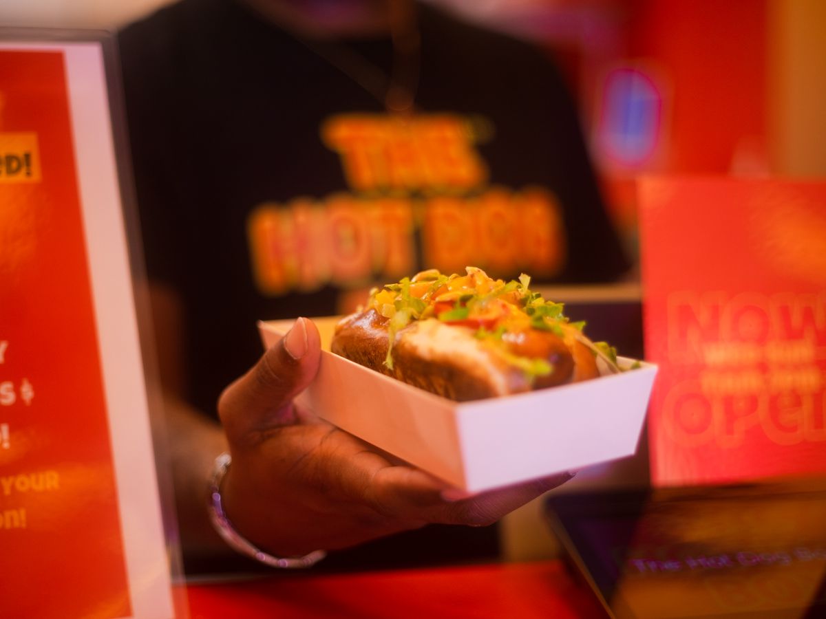 A person’s hand reaches out with a hot dog in a bun.