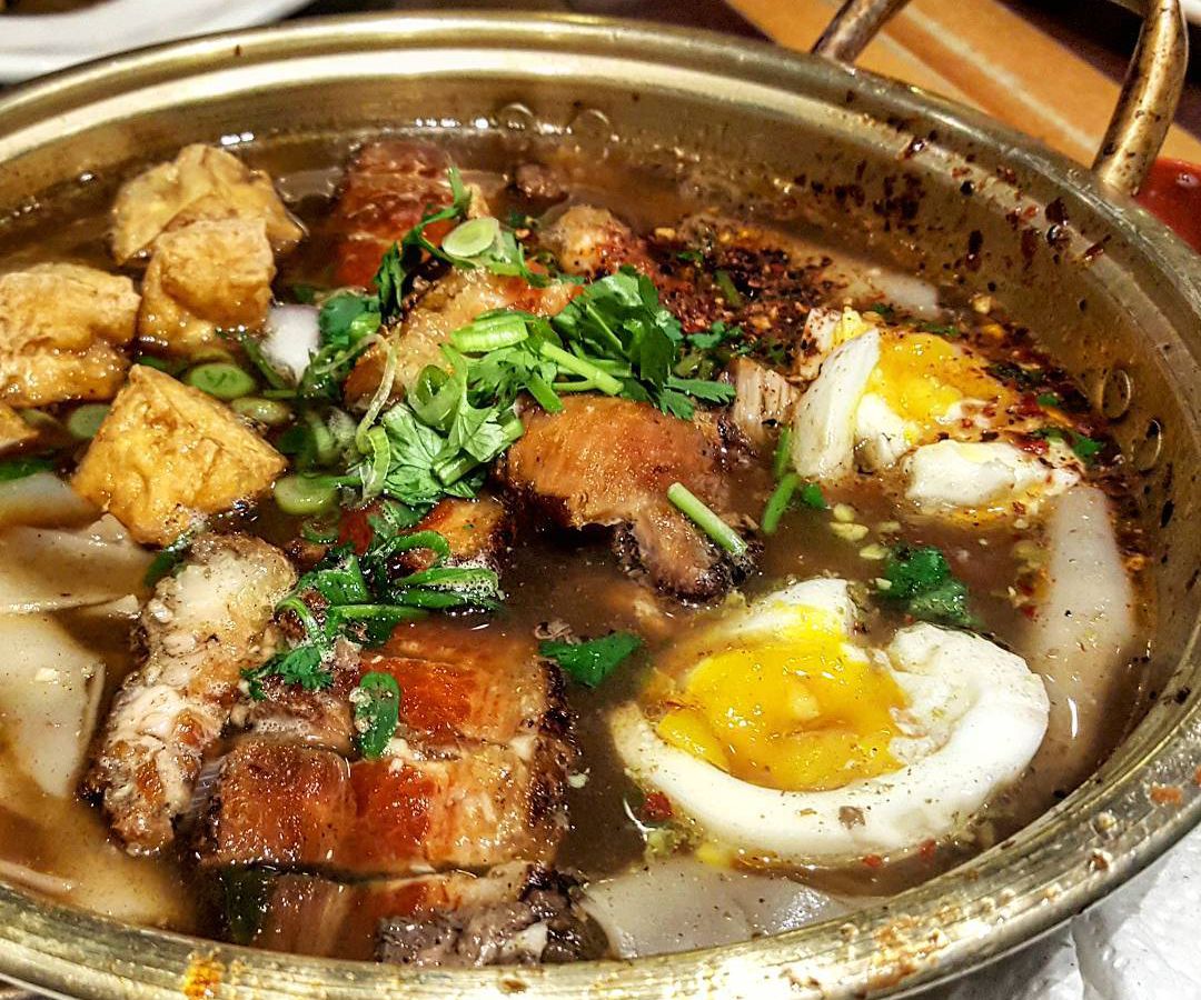 A deep brass bowl is filled with soup. The thin brown broth is packed with fried tofu, a hard-boiled egg, crispy pork belly, and cigar-shaped rolls of noodles.