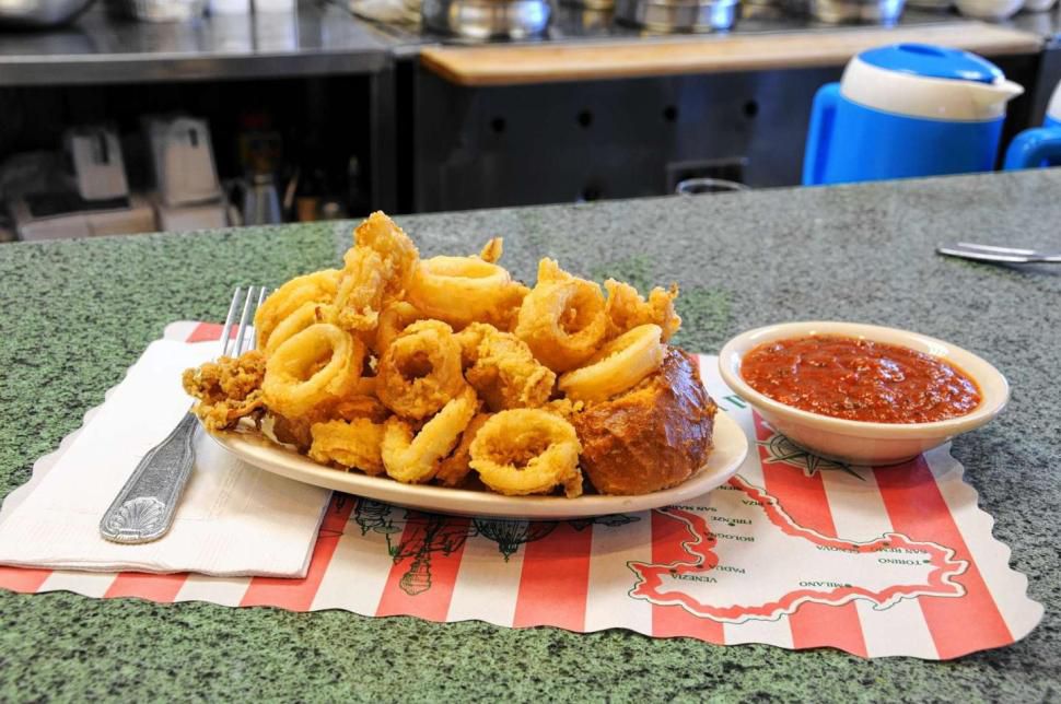 A gingham place mat is topped with a plate teeming with fried calamari, with a white paper napkin with a silver fork on the left, and a small dish with marinara sauce to the right. 