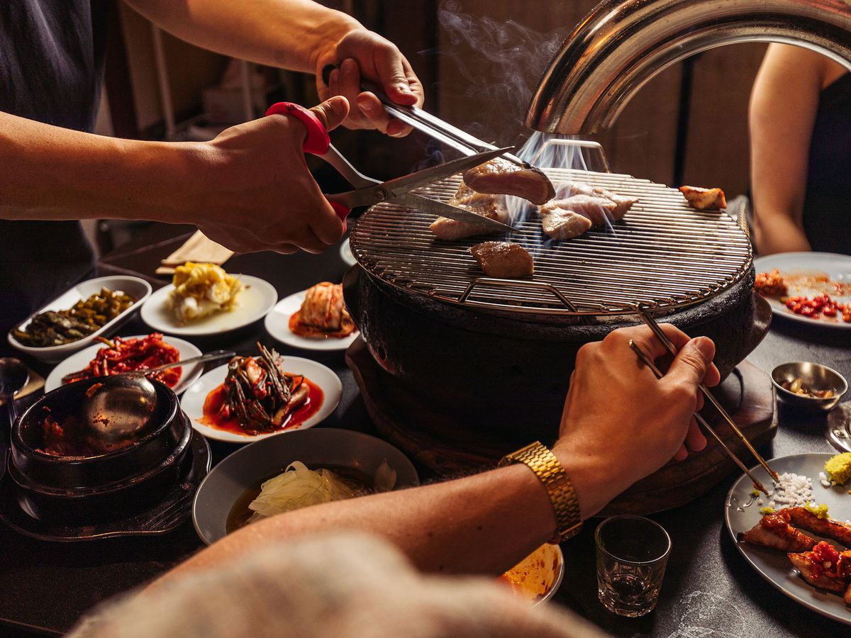 Hands use tongs and scissors to snip a cut of meat over a grill. Other pieces of meat roast on the grill, while diners all around enjoy a table spread with banchan. 