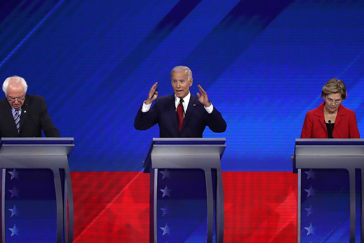Biden, at the center podium, speaks with his hands up, while Sanders to his right and Warren to his left look down at their podiums.