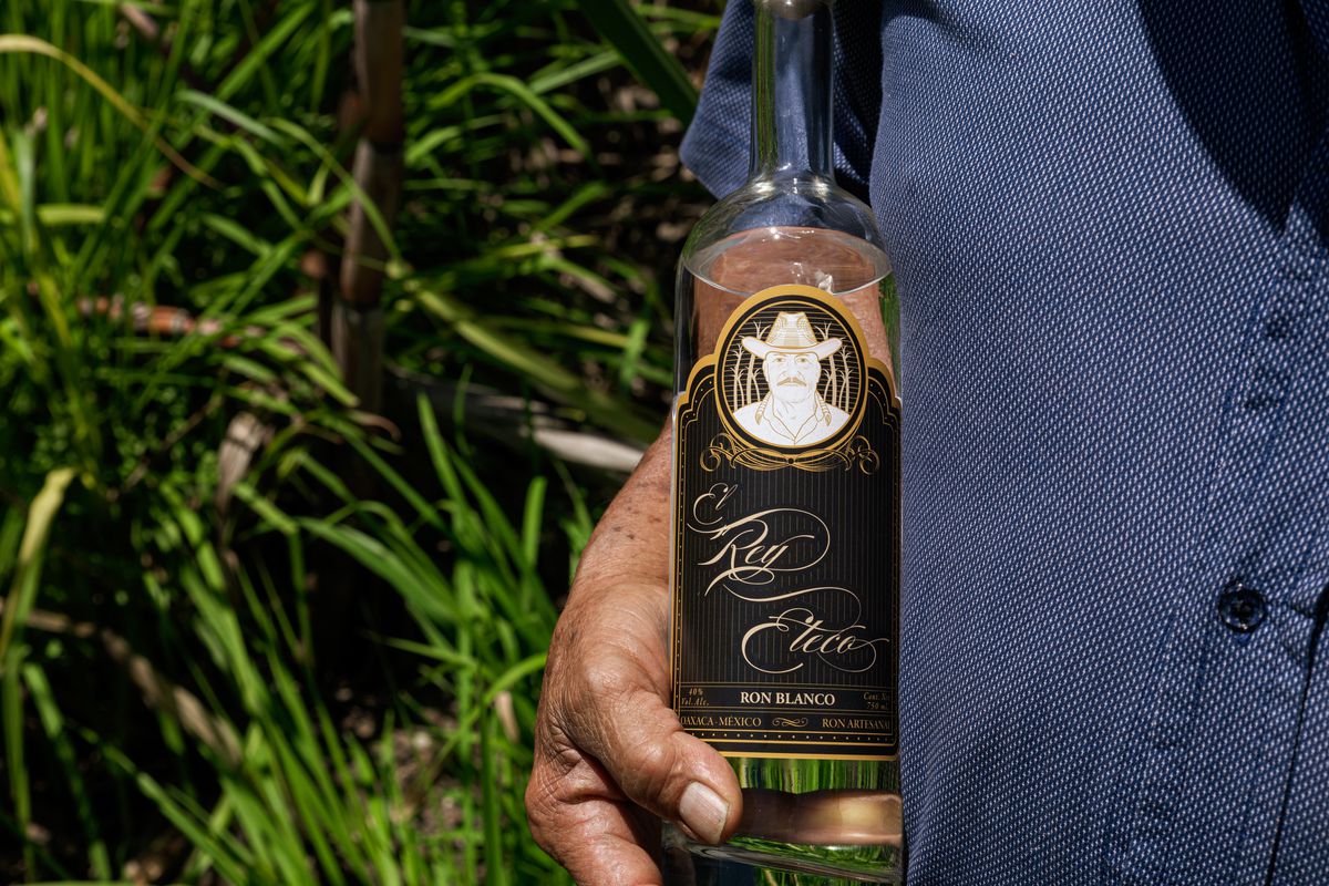 A brown hand holds a bottle of clear rum with the label Rey Eteco. 