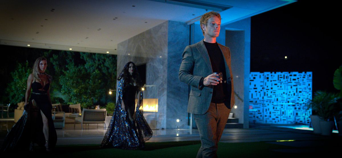 Alfie Allen holds a drink and stands in a mod-looking back yard at night with two other vampires behind him wearing elaborate dresses in Night Teeth