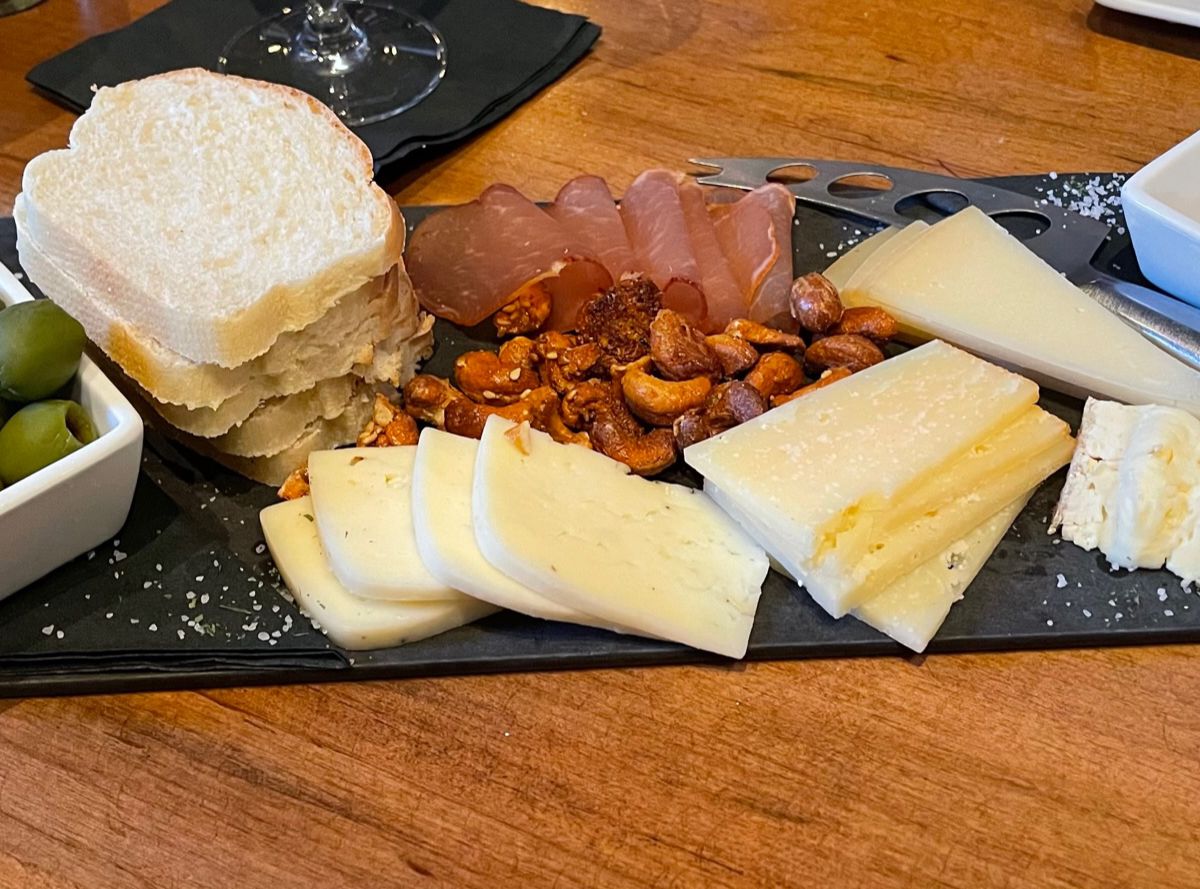 A black slate board topped with various cheeses, ham, bread, and green olives.