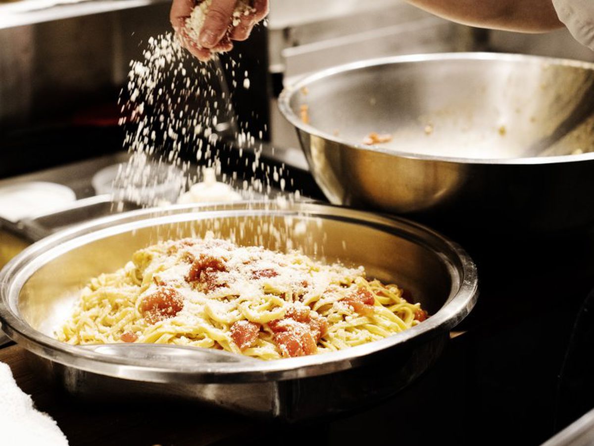 A hand sprinkling parmesan cheese over a large bowl of pasta with tomatoes in it, in a kitchen.