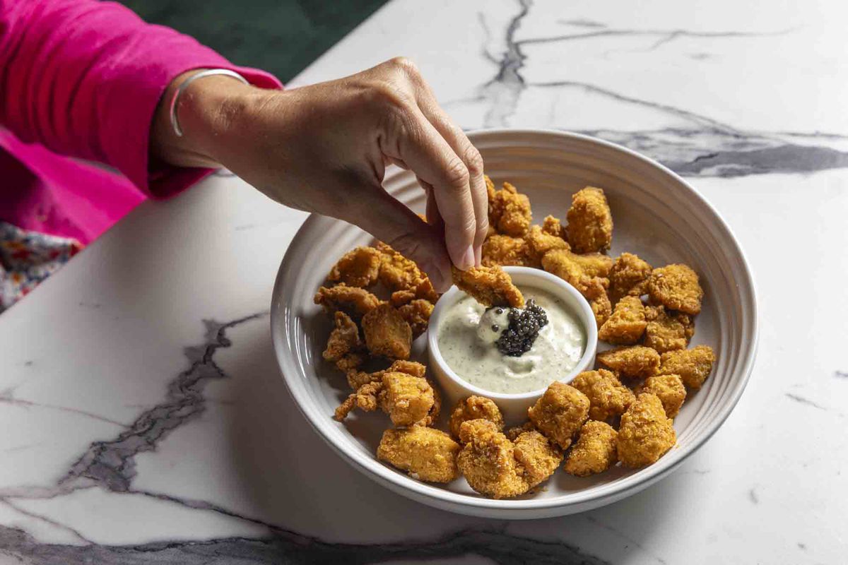 A hand, wearing a pink top, dipping fried chicken into caviar. 