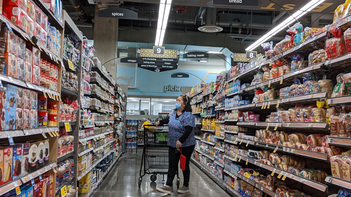 A woman wearing a blue mask stands in the center of an aisle with a shopping cart. 