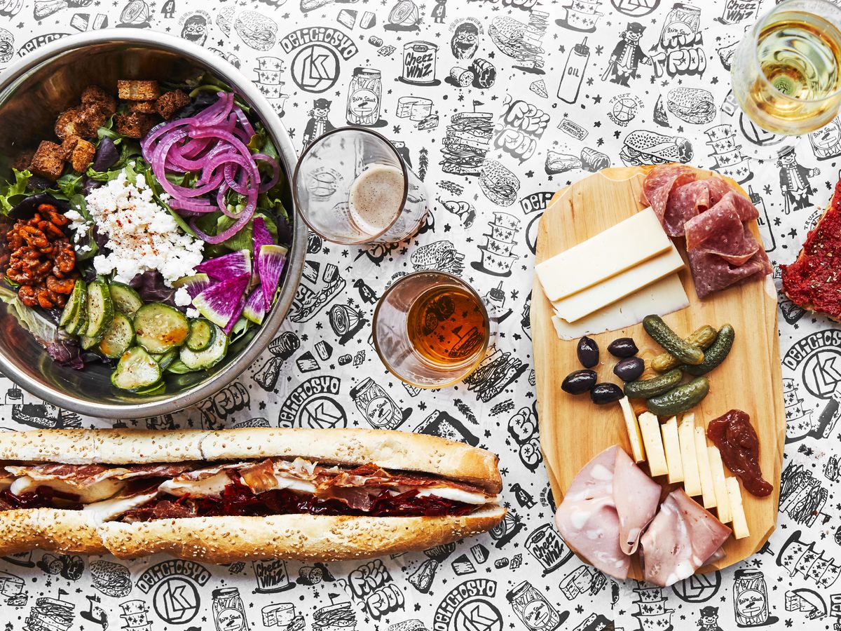 cheeseboard, salad, sandwich on long roll, and glasses of beer and wine on a black and white tablecloth decorated with food images