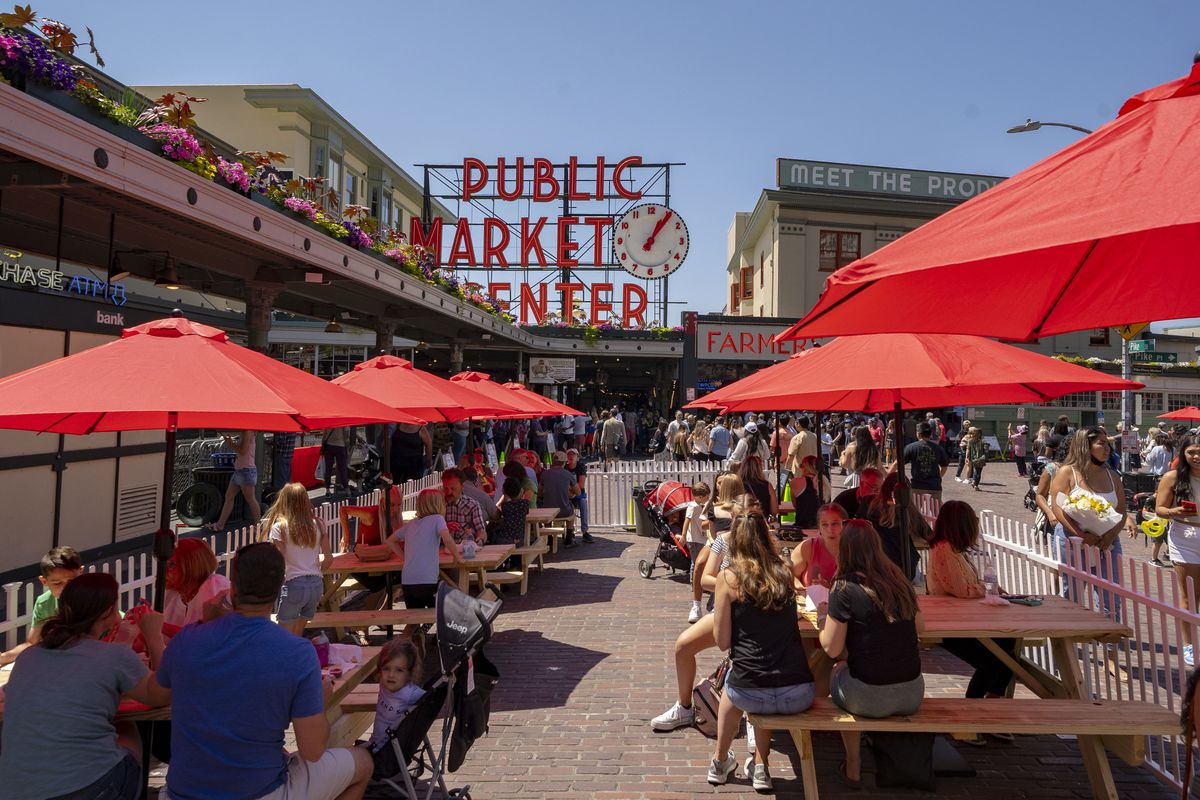 Red umbrellas cover picnic tables on the streets outside Pike Place Market, with the Market’s famed clock and sign in the background.