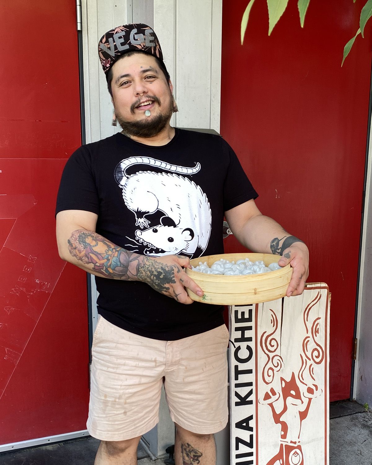 A photo of Cyrus Ichiza holding a steamer of rabbit-shaped bawan dumplings, standing outside the restaurant, in front of a red door and Ichiza Kitchen signage