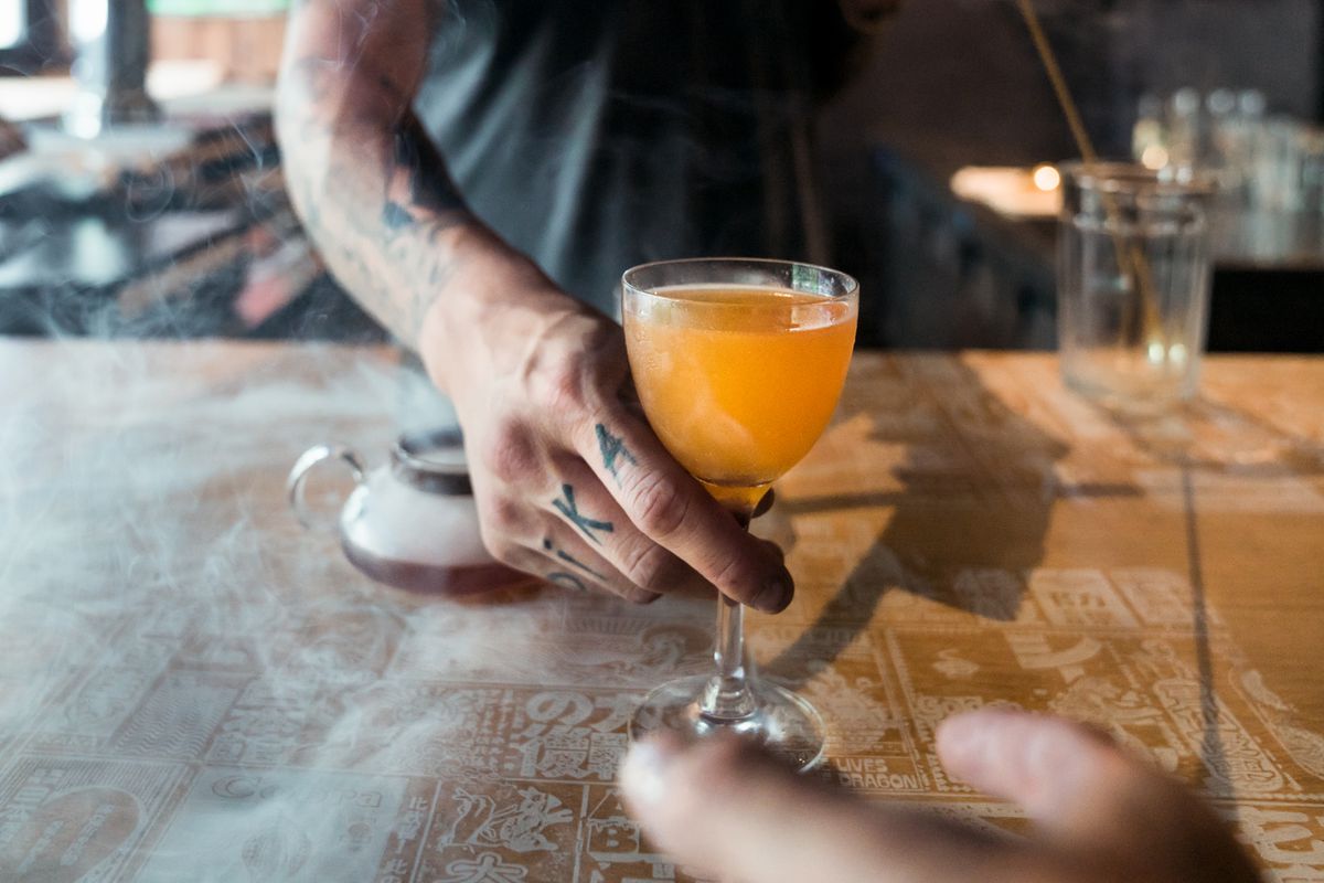 The tattooed arm of a bartender serves up an orange-colored cocktail on a wood bar to a waiting customer.
