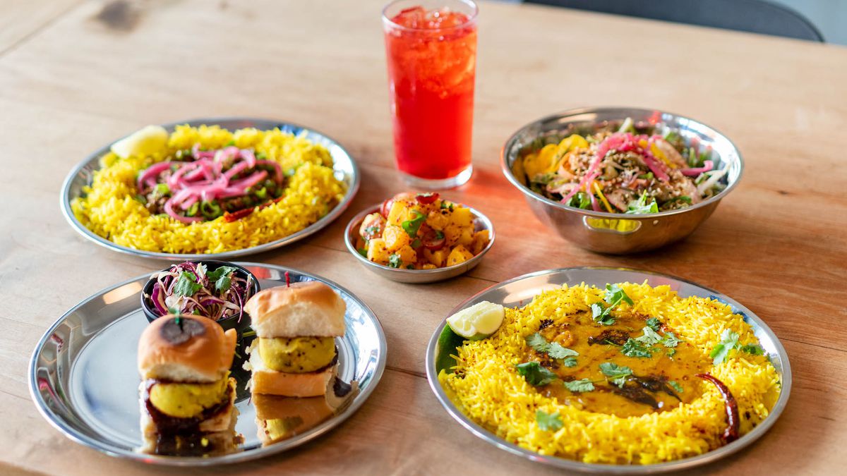 A selection of five silver platters filled with the likes of aloo sliders, kheema, and chana masala, with a light red-colored drink in the center.