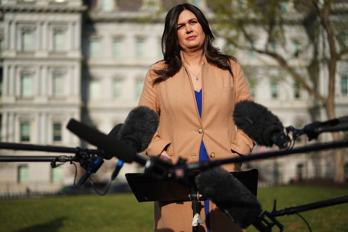 Press Secretary Sarah Sanders with microphones in the foreground and the White House in the background.
