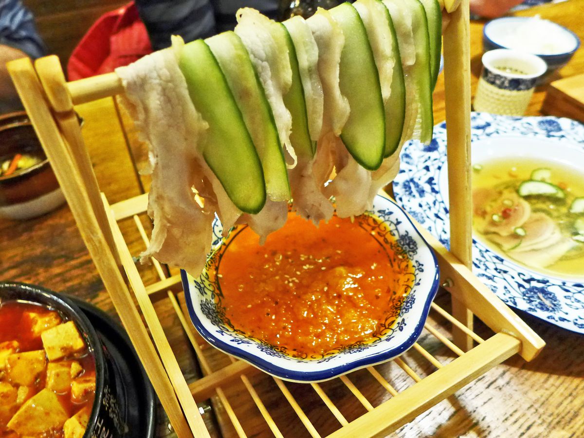 A wooden rack with slices of fatty meat and cucumber hanging over a dish of red chili sauce.