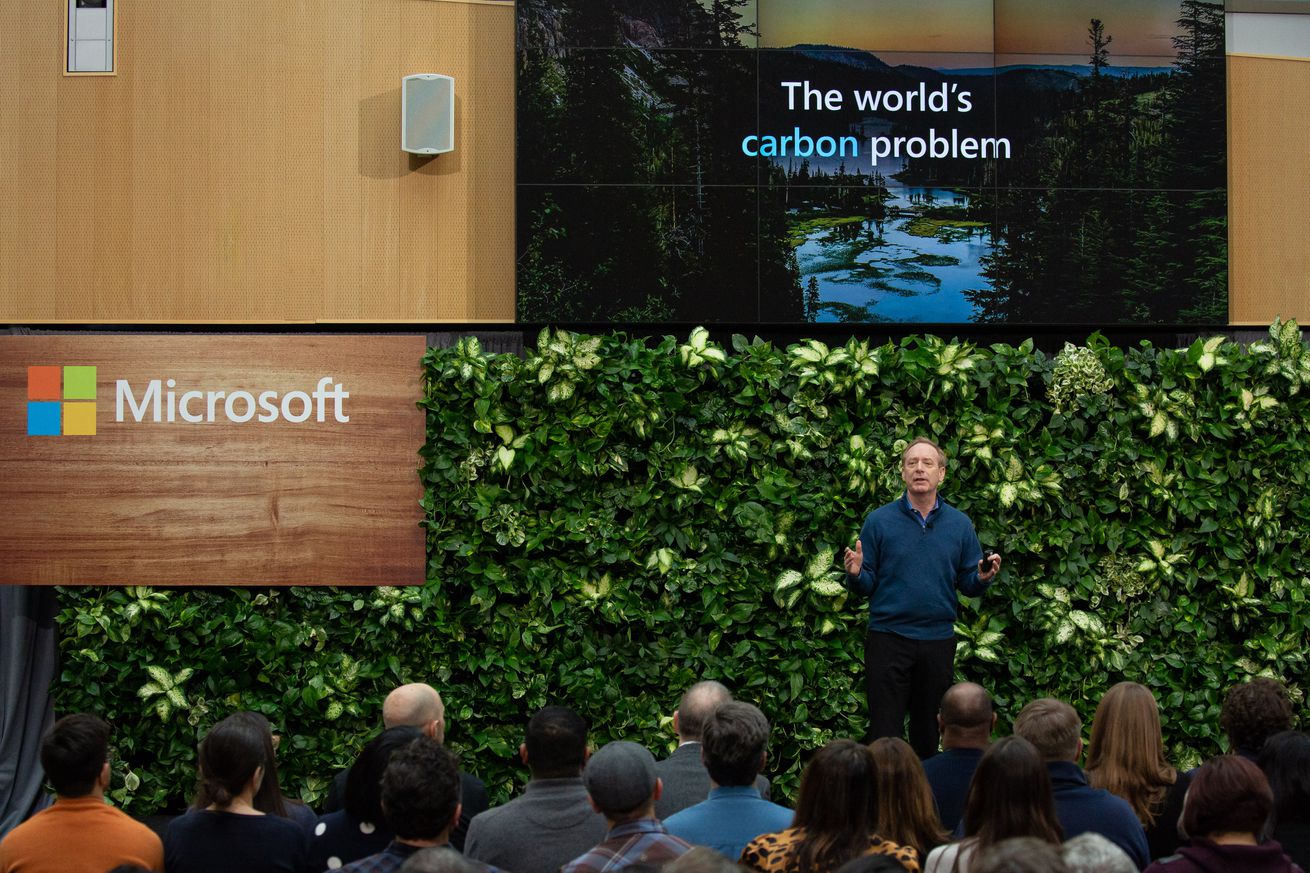 A man speaks to a crowd from a stage. Behind the man is a wall of greenery, the Microsoft logo, and a screen that says “the world’s climate problem.”