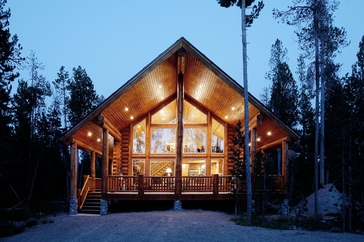 A wooden log cabin with floor to ceiling windows and wooden support beams. The cabin is surrounded by trees. 