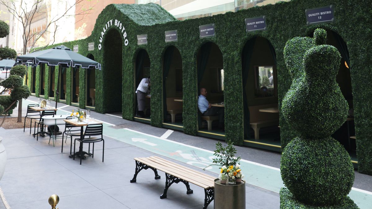 A row of outdoor seating booths covered in greenery on the side of the road with benches and tables set up on the sidewalk facing the road