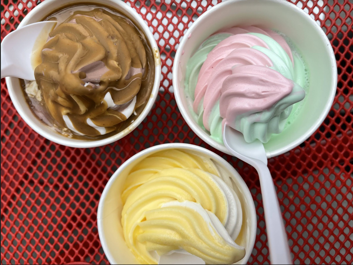 A red grated table features three white cups filled with brown, pink-and-green, and yellow-and-white soft serve.