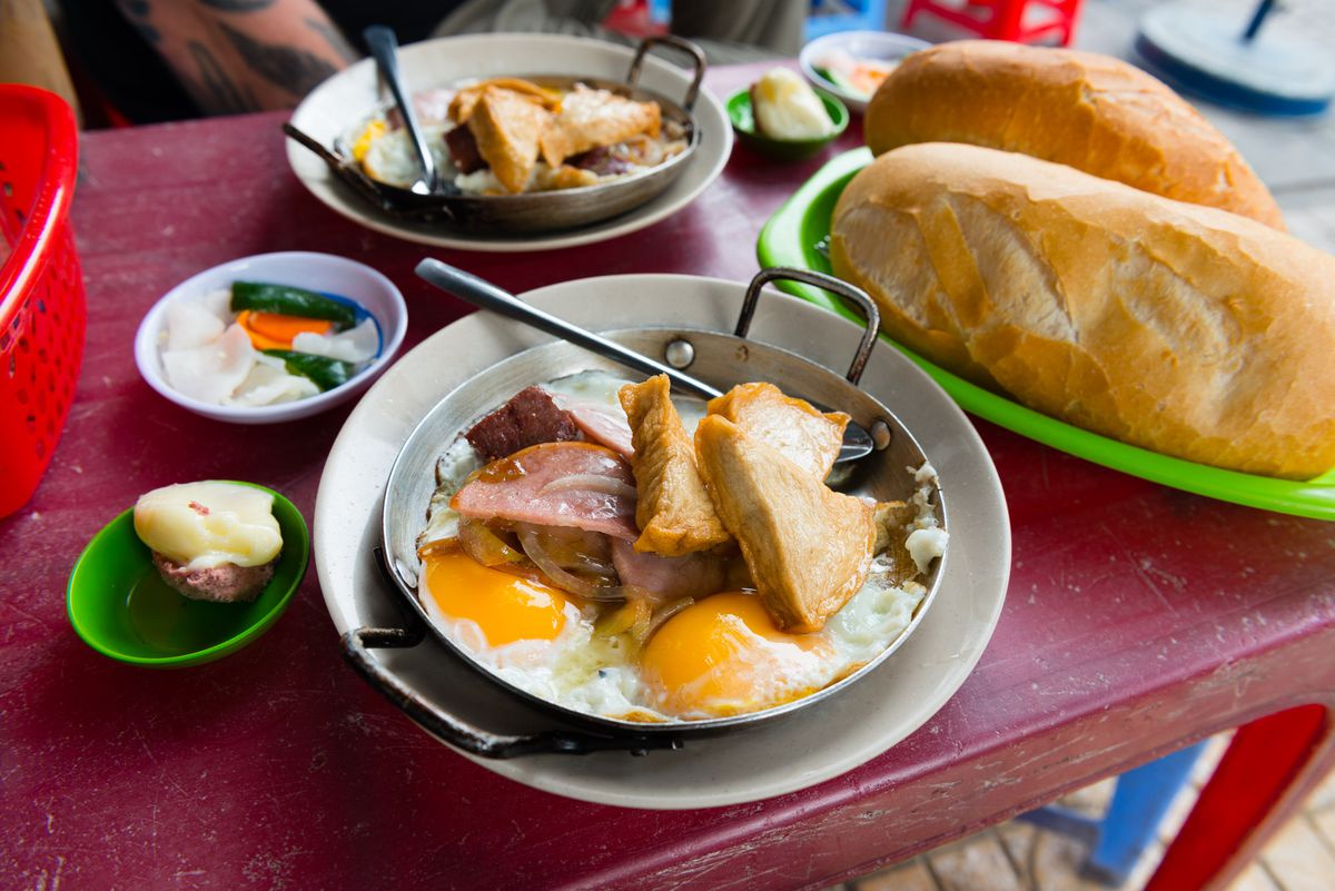 A plate of eggs topped with slabs of meat and fried tofu, served with large loaves of bread.
