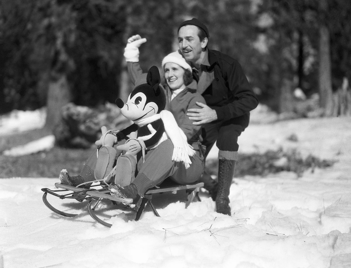 In an undated black-and-white portrait, Walk Disney crouches behind his wife Lillian and a Mickey Mouse doll, both sitting on an old-fashioned wooden sled with metal runners, outdoors in the snow