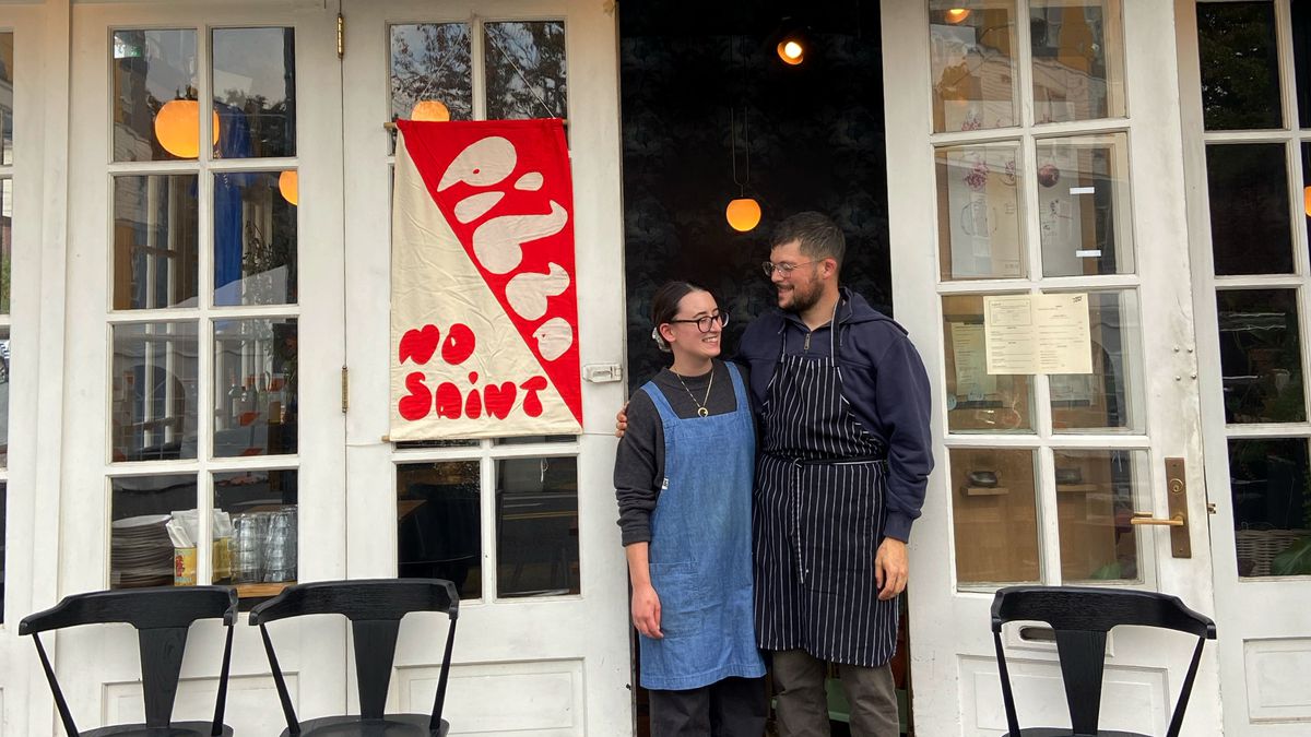 Two people stand outside a restaurant next to a banner that reads “pizza” and “No Saint.”